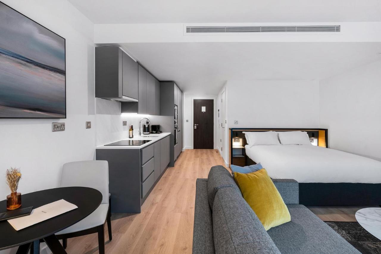 Citysuites 2 Aparthotel Manchester Esterno foto The photo shows a modern and stylish studio apartment. It features a kitchenette with dark cabinetry and a small dining table with two chairs. There's a cozy sofa with colorful cushions, positioned next to the table. In the background, a large bed wi