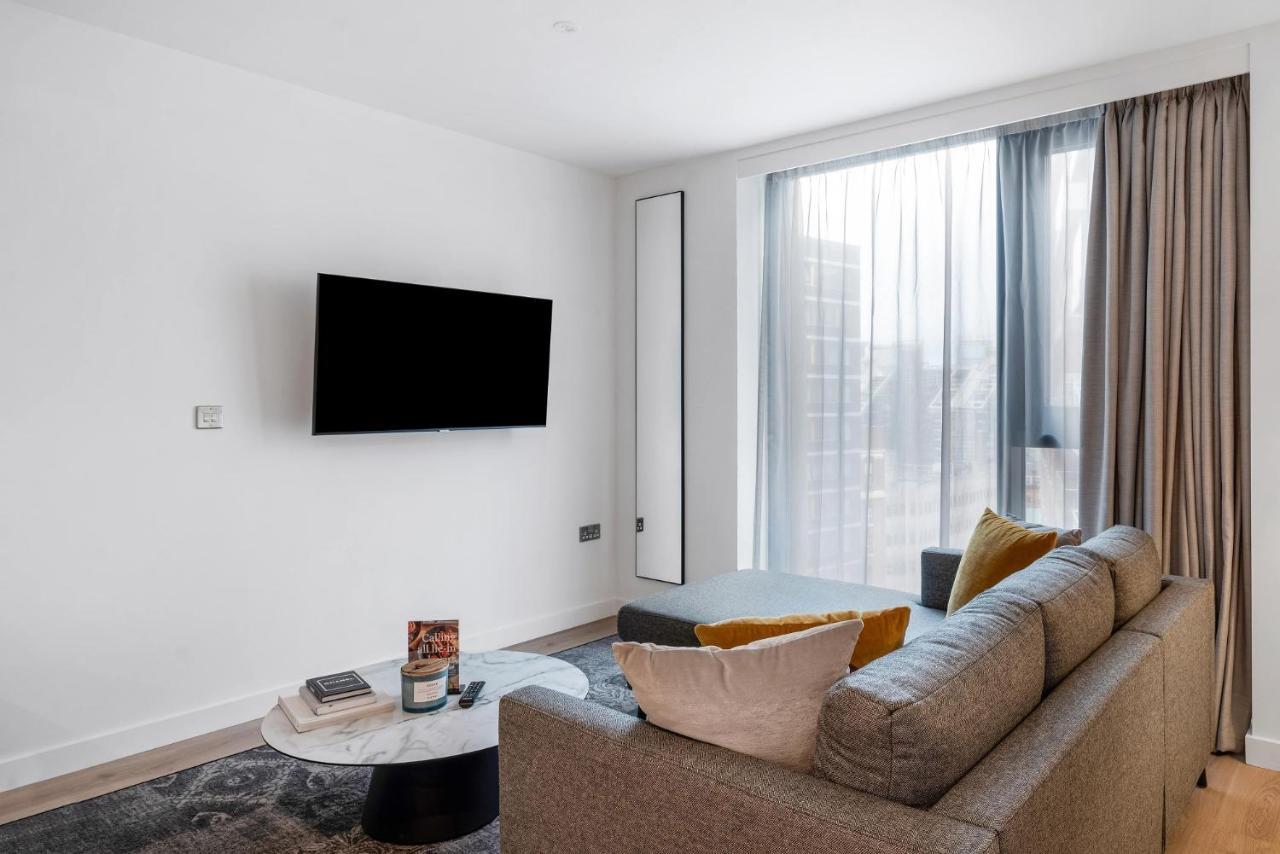 Citysuites 2 Aparthotel Manchester Esterno foto The image shows a modern living room with a minimalist design. There is a large flat-screen TV mounted on the white wall. A comfortable gray sofa with textured cushions occupies the center of the room, complemented by a round coffee table in front of
