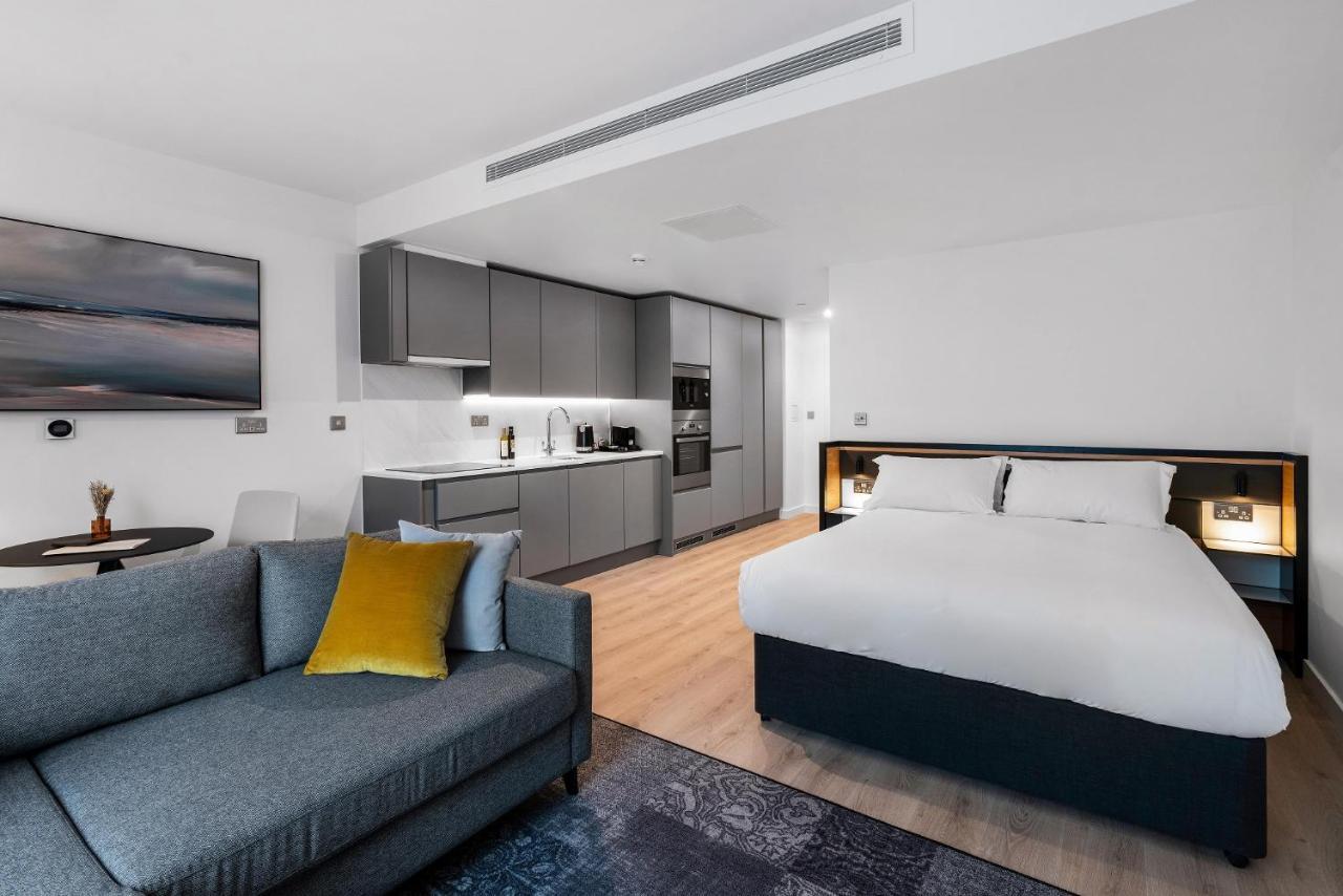 Citysuites 2 Aparthotel Manchester Esterno foto The photo shows a modern studio apartment. On one side, there is a comfortable gray sofa adorned with a yellow cushion. Nearby, a round table is placed, likely intended for dining or casual sitting. 

The kitchen area features sleek gray cabinetry wi