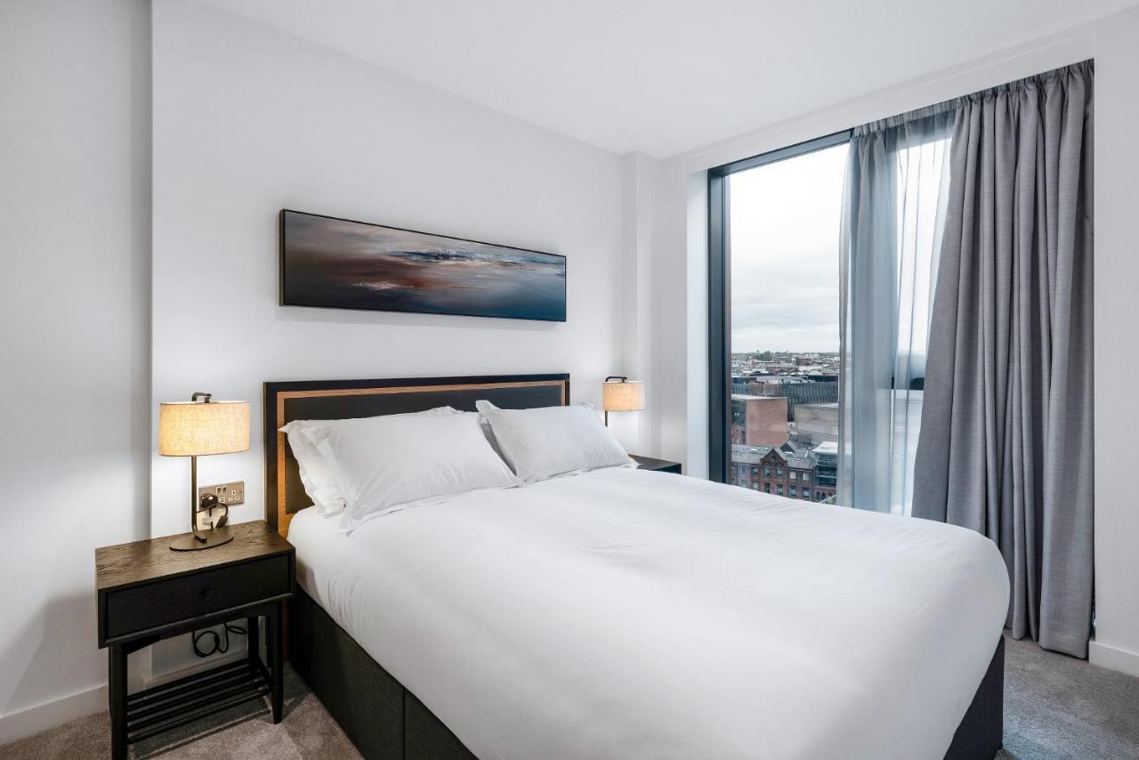 Citysuites 2 Aparthotel Manchester Esterno foto The image shows a modern bedroom featuring a large bed with a white duvet and two pillows. On either side of the bed are small nightstands with lamps. A large window occupies one wall, draped with light gray curtains, allowing natural light to enter 