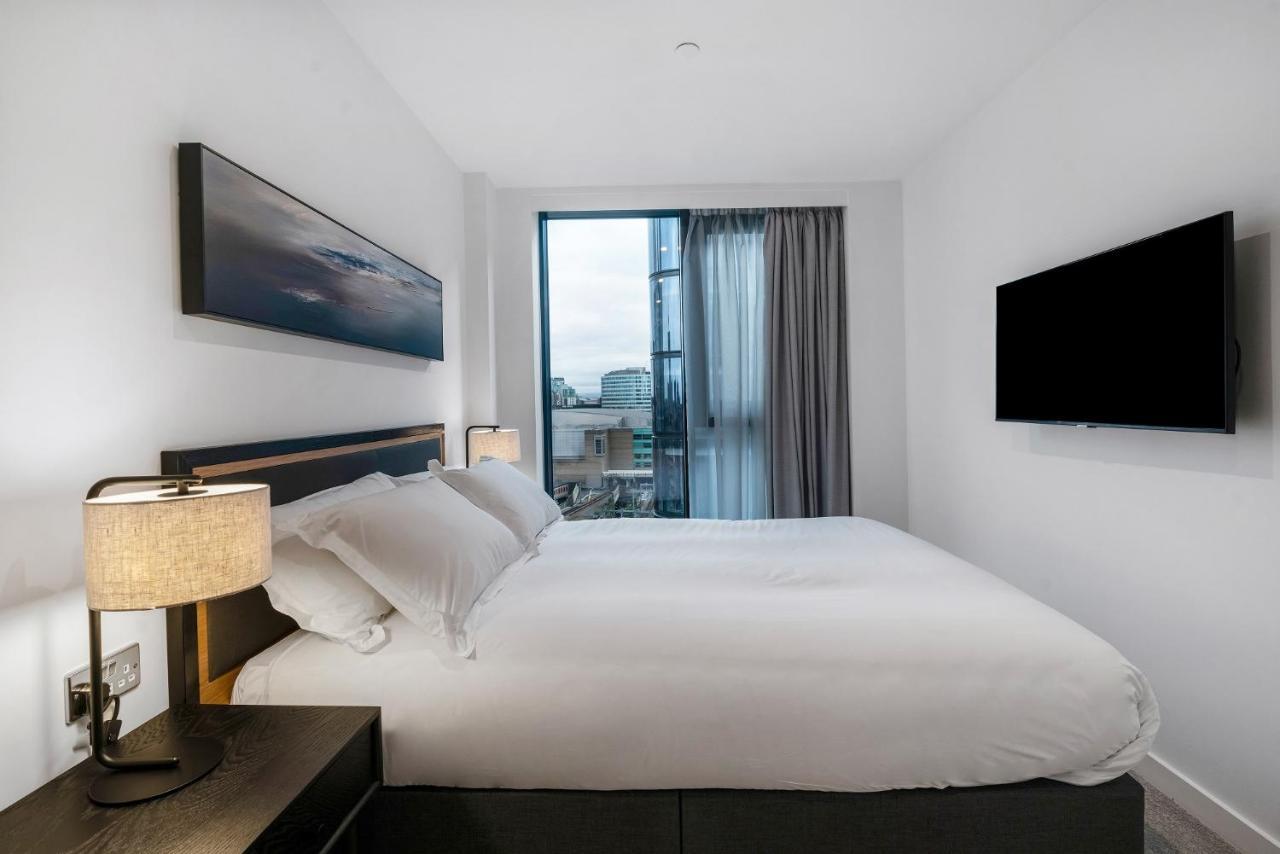 Citysuites 2 Aparthotel Manchester Esterno foto The photo shows a modern bedroom with a minimalist aesthetic. It features a large bed with white bedding and two pillows. On either side of the bed, there are bedside tables with lamps. A large window lets in natural light, covered by sheer curtains.