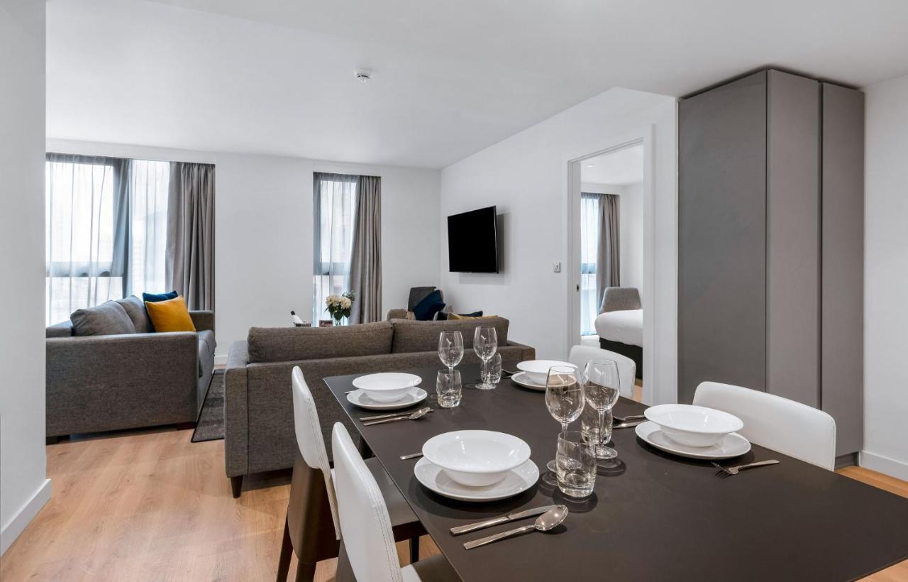 Citysuites 2 Aparthotel Salford  Esterno foto The photo shows a modern living space with an open layout. In the foreground, there is a dining table set for a meal, featuring multiple plates, glasses, and cutlery arranged neatly. In the background, there is a comfortable-looking gray sofa with a 