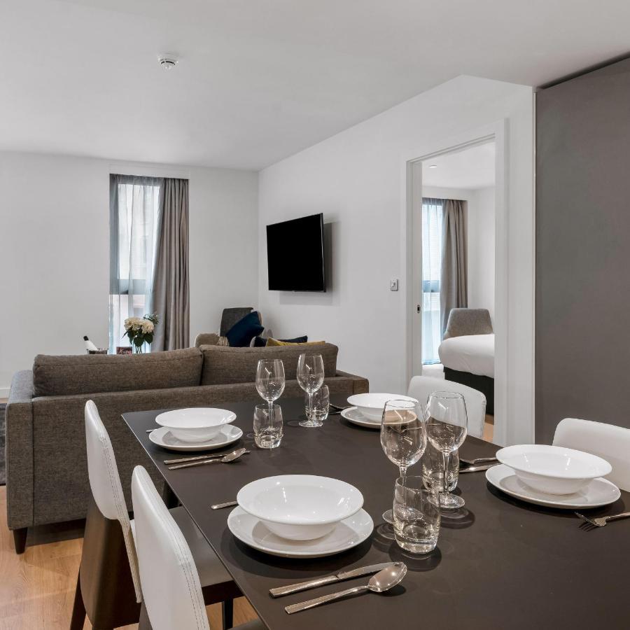 Citysuites 2 Aparthotel Manchester Esterno foto The photo shows a modern, well-lit living space that features a dining area and a sitting area. In the foreground, there is a dining table set for six, with white plates and crystal glasses arranged neatly. The table is surrounded by white chairs. 

