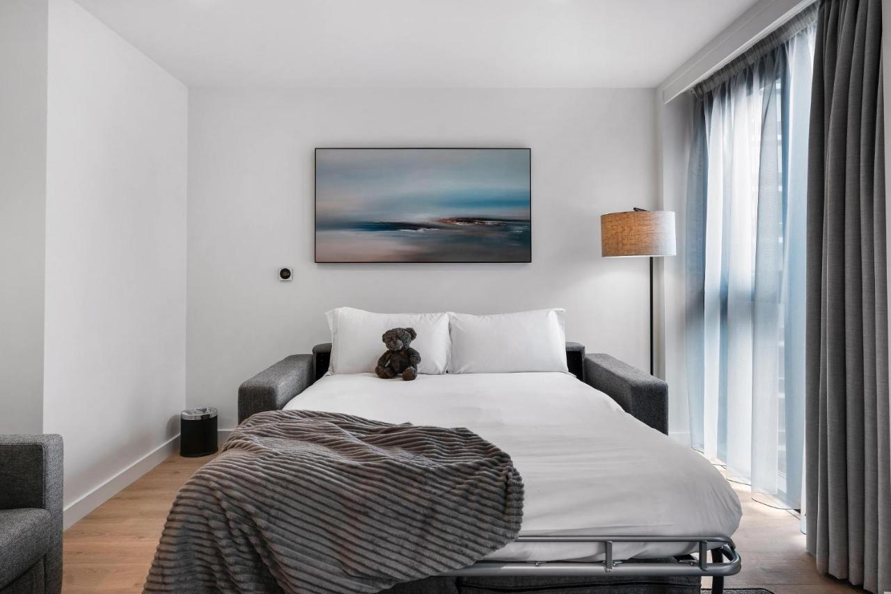 Citysuites 2 Aparthotel Manchester Esterno foto The photo shows a modern bedroom featuring a bed with white bedding and a decorative throw blanket. There is a teddy bear placed on the bed. A piece of abstract art is hanging on the wall above the bed. The room has a large window with sheer curtains