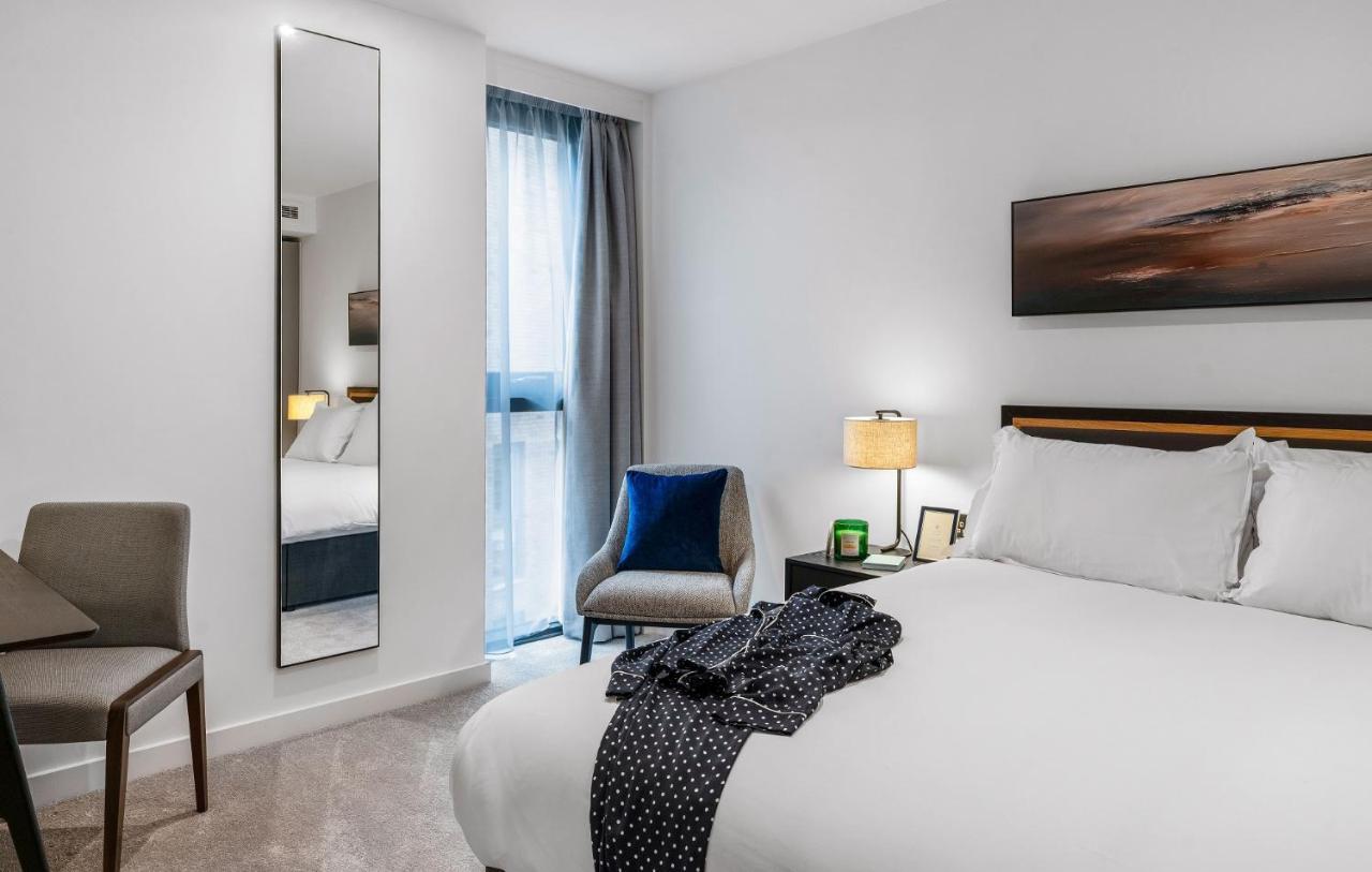 Citysuites 2 Aparthotel Manchester Esterno foto The photo shows a modern hotel room. It features a neatly made bed with white linens and a dark-colored throw draped over it. There is a small bedside table with a lamp next to the bed. A comfortable chair in a light color is positioned near the wind