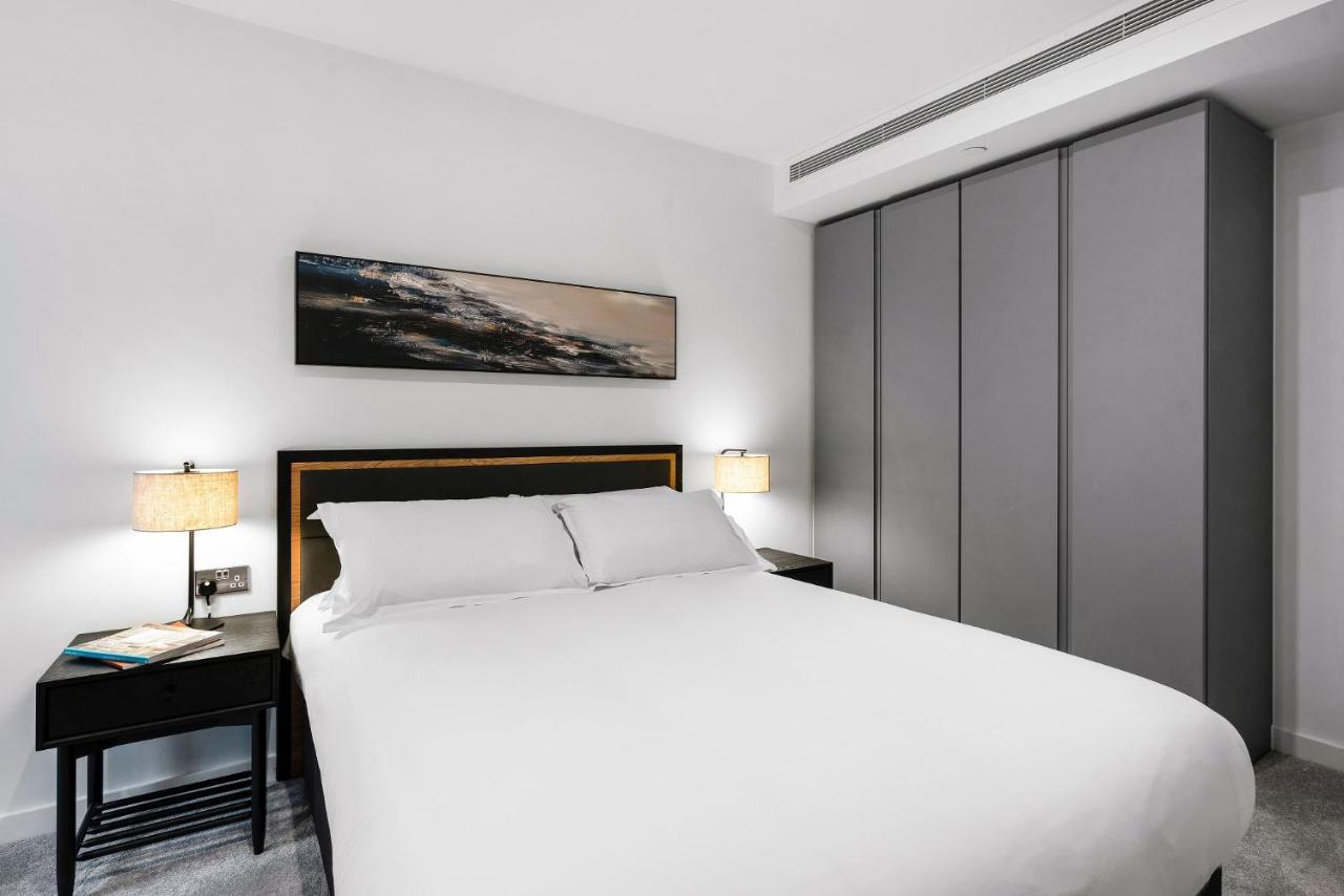 Citysuites 2 Aparthotel Manchester Esterno foto The photo shows a modern bedroom designed with a minimalist aesthetic. The focal point is a large bed with white bedding, positioned against a wall. On either side of the bed, there are black nightstands topped with lamps, providing a warm glow. Abov