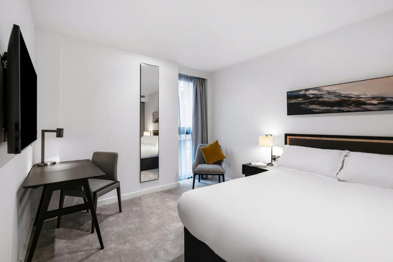 Citysuites 2 Aparthotel Manchester Esterno foto The photo shows a modern hotel room with a clean and minimalist design. It features a large bed with white linens, a small desk with a chair, and a mirror mounted on the wall. There is a chair in one corner with a decorative pillow, and the room has 