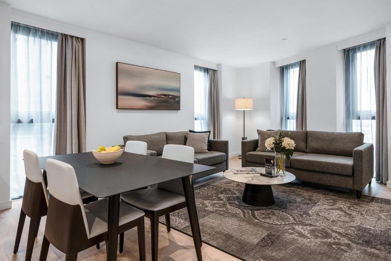 Citysuites 2 Aparthotel Manchester Esterno foto The photo depicts a modern and stylish interior of a living space. On the left, there is a dining table with four chairs, featuring a bowl of fruits on the table. In the background, there's a cozy seating area with a gray sofa and a smaller round cof