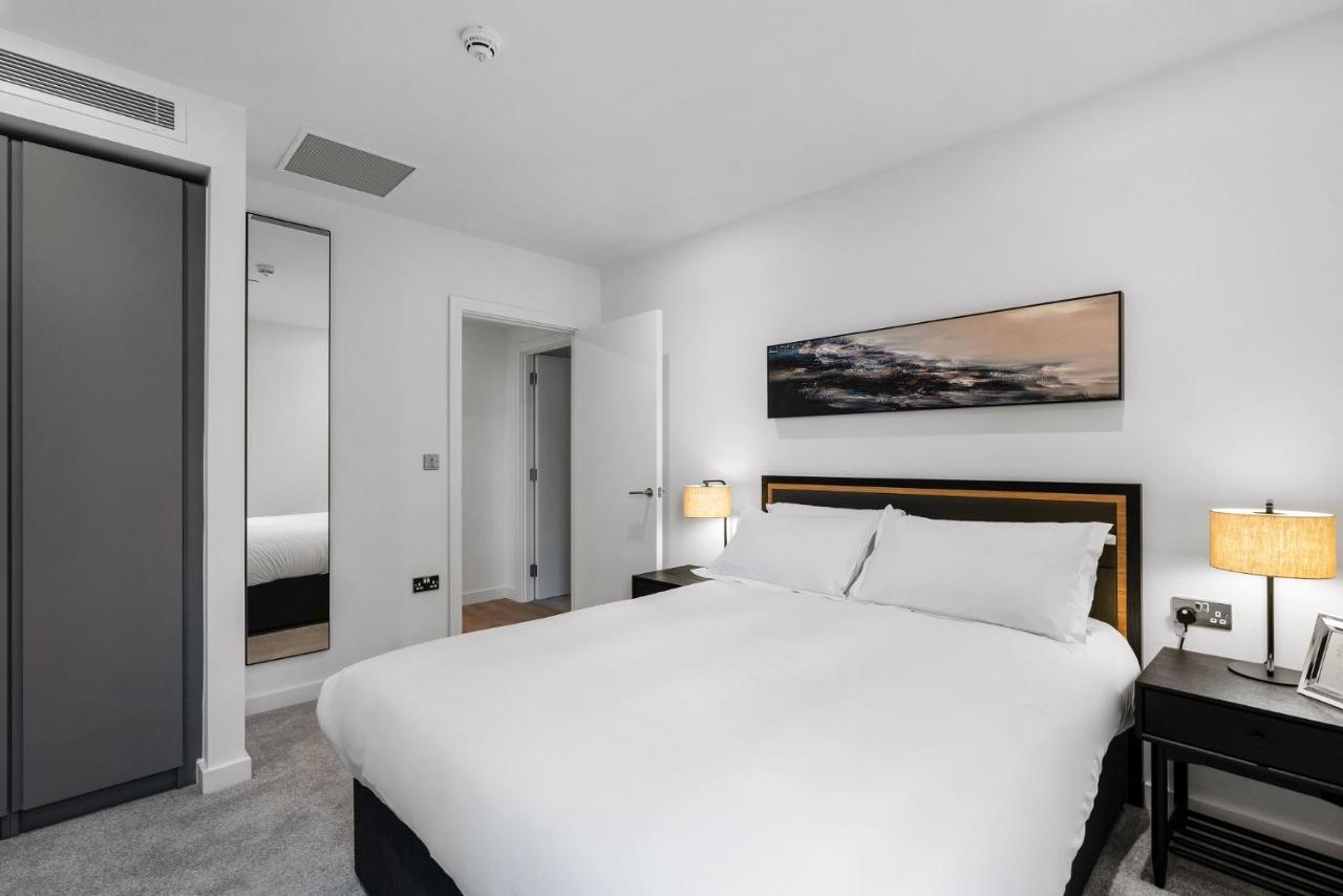 Citysuites 2 Aparthotel Salford  Esterno foto The photo shows a modern bedroom featuring a large double bed with white bedding. There are two bedside tables with lamps on either side of the bed. The walls are painted white, and there's a piece of artwork above the bed that depicts a landscape. A