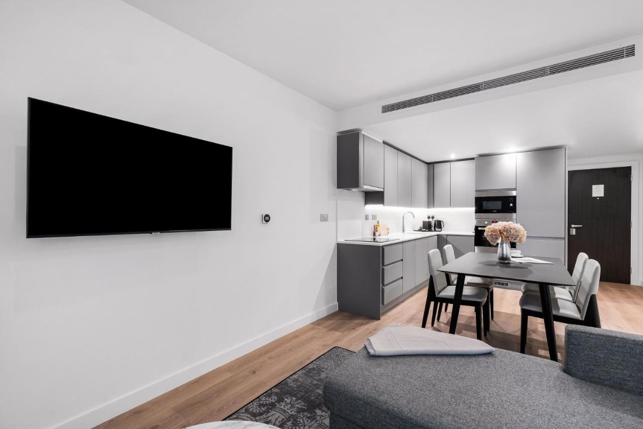 Citysuites 2 Aparthotel Manchester Esterno foto The photo shows a modern, minimalist living space. On the left, there is a large flat-screen TV mounted on the wall. To the right, a kitchen area features sleek gray cabinets and appliances, including a built-in microwave. In front of the kitchen, th