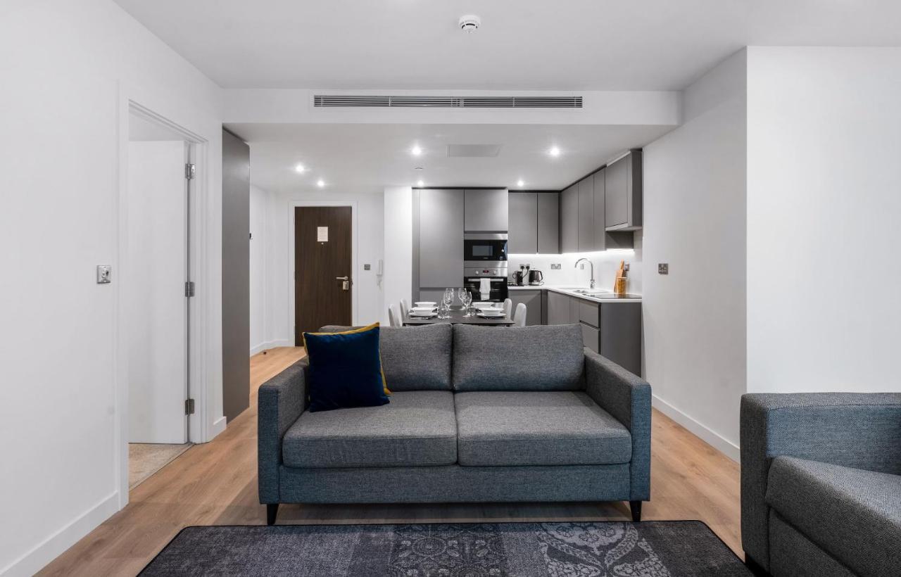 Citysuites 2 Aparthotel Manchester Esterno foto The photo shows a modern interior space, likely a living area or apartment. In the foreground, there is a gray sofa with a blue cushion positioned on a textured rug. To the back, there is an open kitchen with sleek cabinetry and appliances, visible a