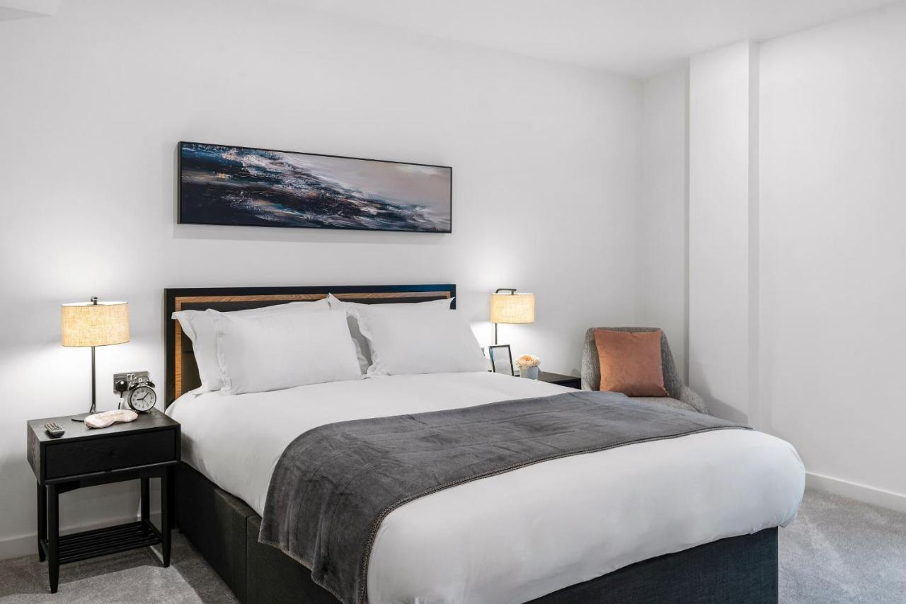 Citysuites 2 Aparthotel Manchester Esterno foto The photo shows a modern bedroom featuring a large bed with crisp white linens and a grey throw blanket. On either side of the bed, there are bedside tables with lamps. A piece of abstract art hangs above the headboard, and there is a chair in a corn