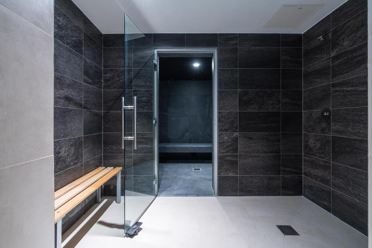 Citysuites 2 Aparthotel Manchester Esterno foto The photo shows a modern, minimalist shower area. The space is predominantly tiled with dark gray tiles on the walls, giving it a sleek and sophisticated look. There is a glass door that leads to another room, and a wooden bench is positioned against