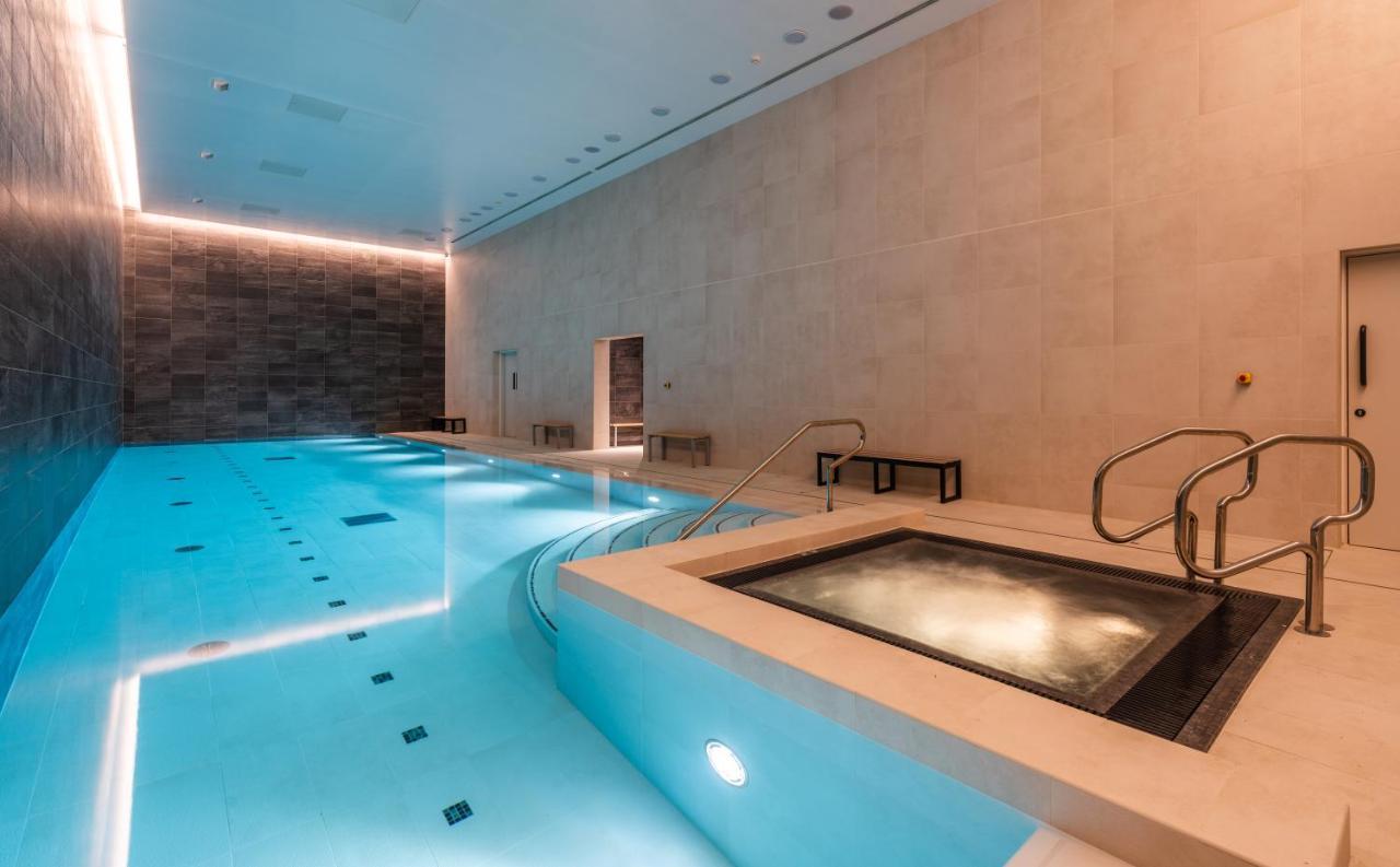 Citysuites 2 Aparthotel Manchester Esterno foto The photo shows an indoor swimming pool area. The pool is a bright blue color and features smooth, tiled surfaces. On one side, there is a hot tub area with steps leading into it. The walls are made of light-colored stone, and there is ambient lighti