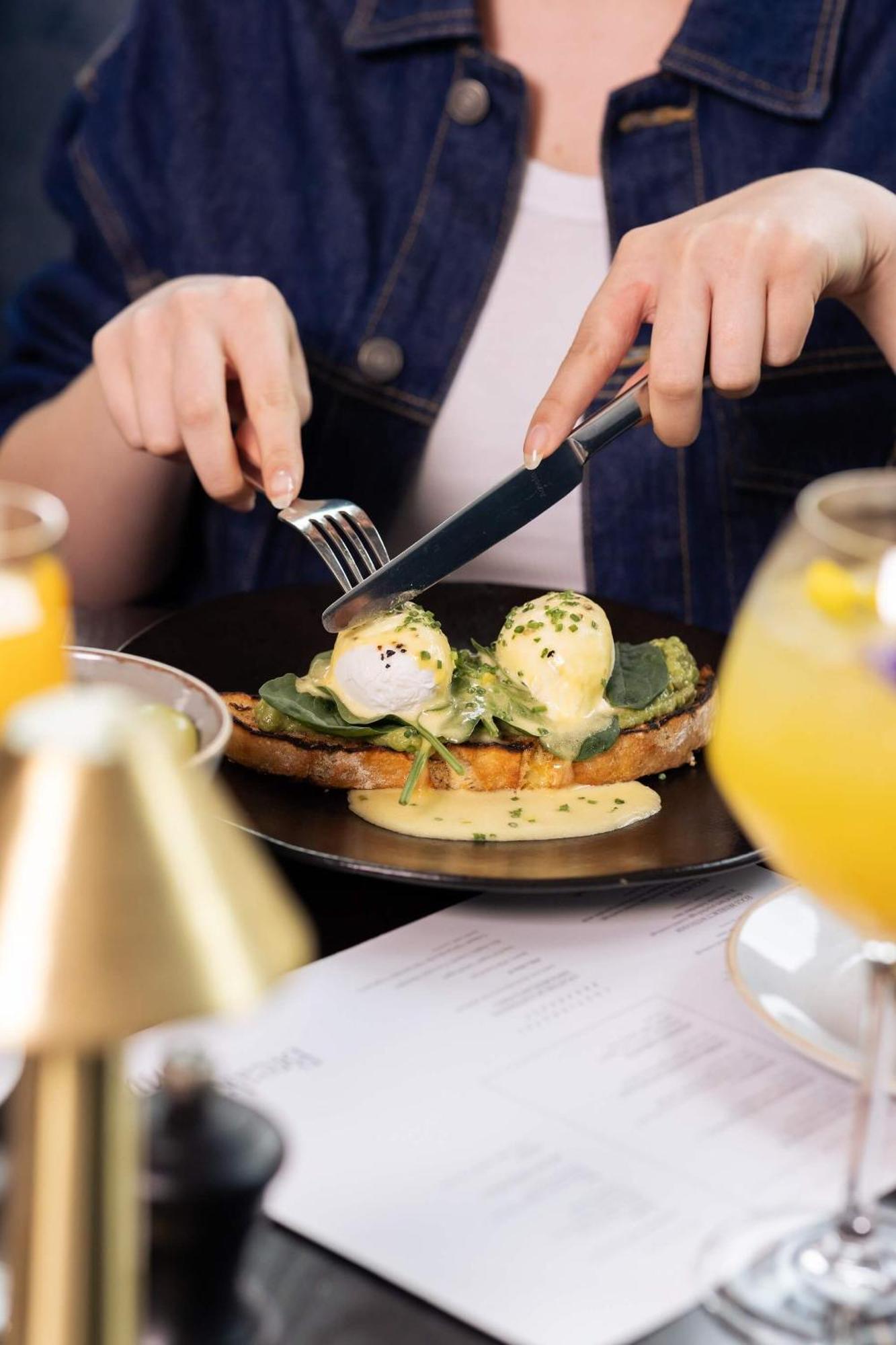 Citysuites 2 Aparthotel Manchester Esterno foto The photo shows a person preparing to eat a dish of eggs Benedict. They are holding a knife and fork, ready to cut into the meal, which features poached eggs on top of a slice of toast, accompanied by spinach and a creamy hollandaise sauce drizzled o