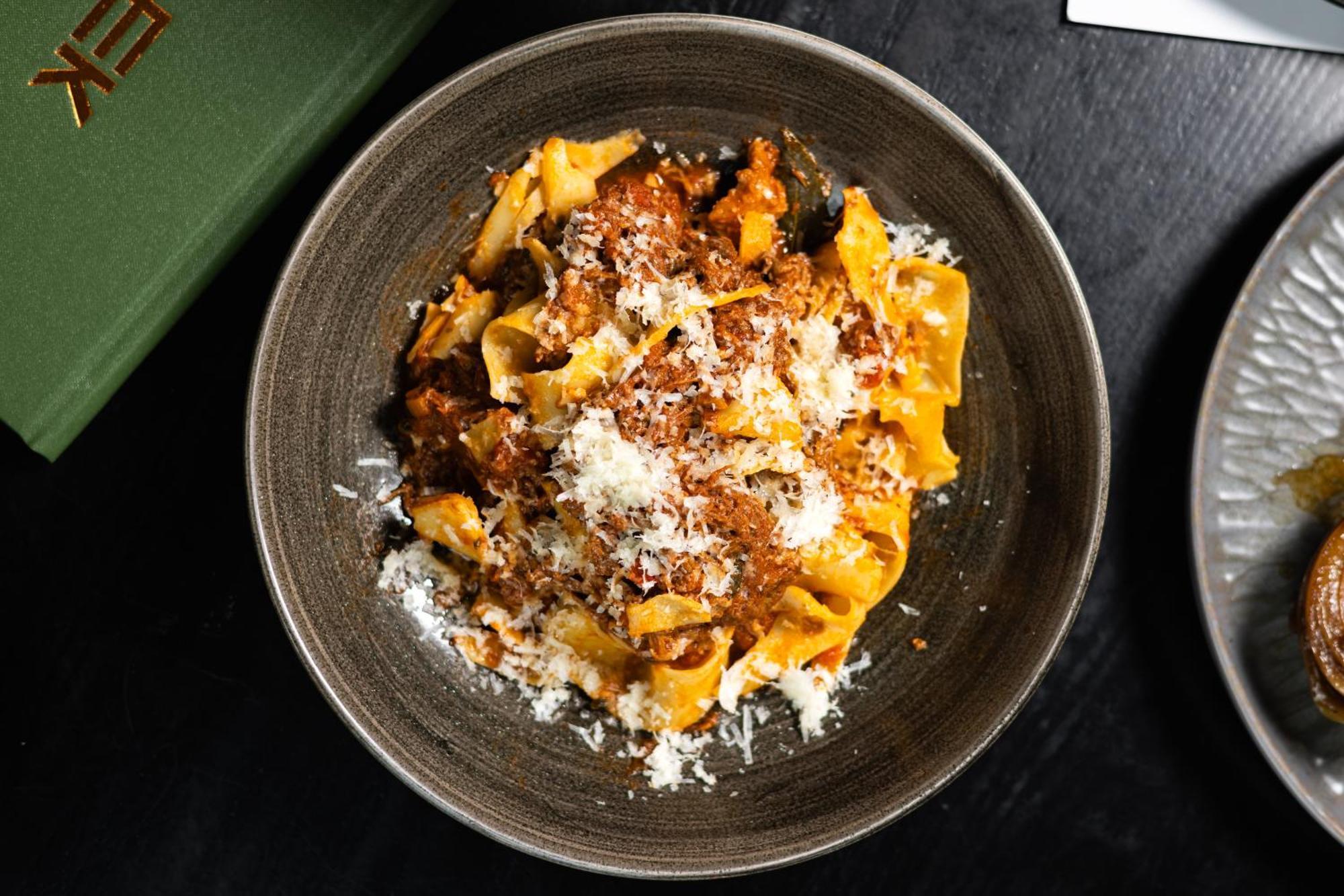 Citysuites 2 Aparthotel Manchester Esterno foto The photo shows a bowl of pasta featuring wide, ribbon-like noodles, likely pappardelle, fully coated in a rich meat sauce. The dish is garnished with a generous sprinkling of grated cheese, adding a creamy texture to the presentation. The dark bowl 